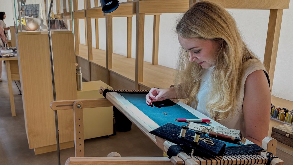 A woman with long blonde hair leans over a work station as she embroiders on a large teal fabric.