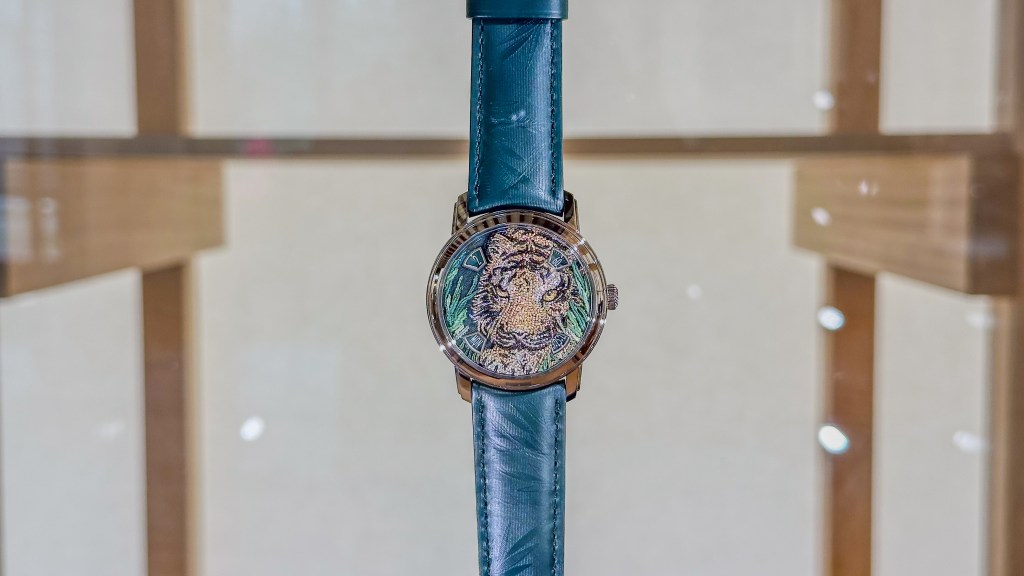 A detail of a watch with a teal band and an intricately embroidered dial of a lion's face.