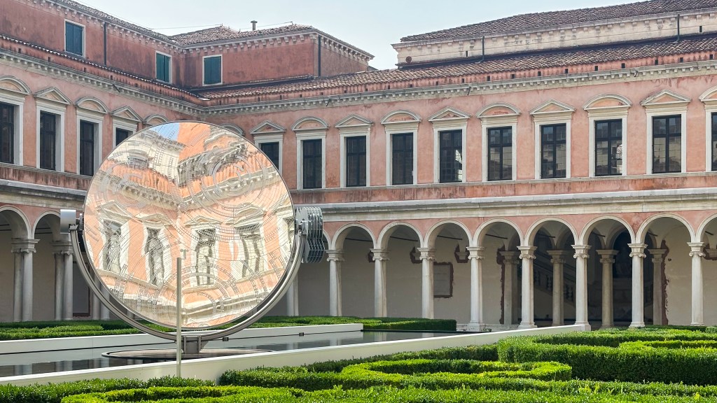 The cloister of a Renaissance building with a circular mirror sculpture in the middle, reflecting another side of the building.