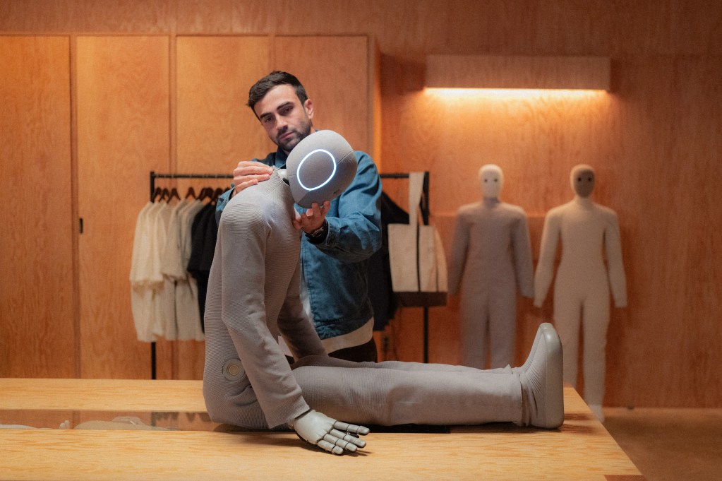 Dar Sleeper holds a 1X NEO robot upright in a light wood room displaying clothing and more NEOS.