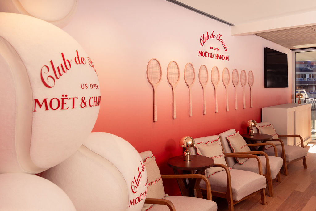 A view of the Moët & Chandon suite at US Open with oversized tennis balls in a corner and wooden rackets hanging on a red and white gradient wall.