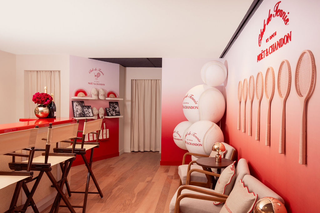 A view of the Moët & Chandon suite at US Open with oversized tennis balls in a corner, archival tennis champion photographs, wooden rackets and a red and white gradient wall.