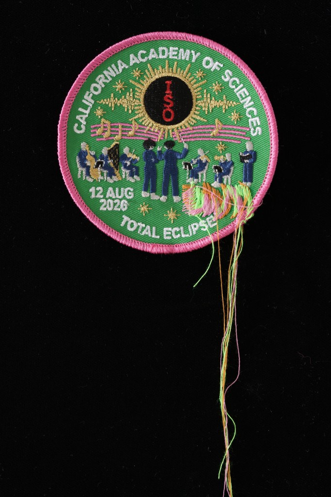 A green and pink patch that says California Academy of Sciences and Total Eclipse on it with figures of conductors and musicians embroidered on it. 