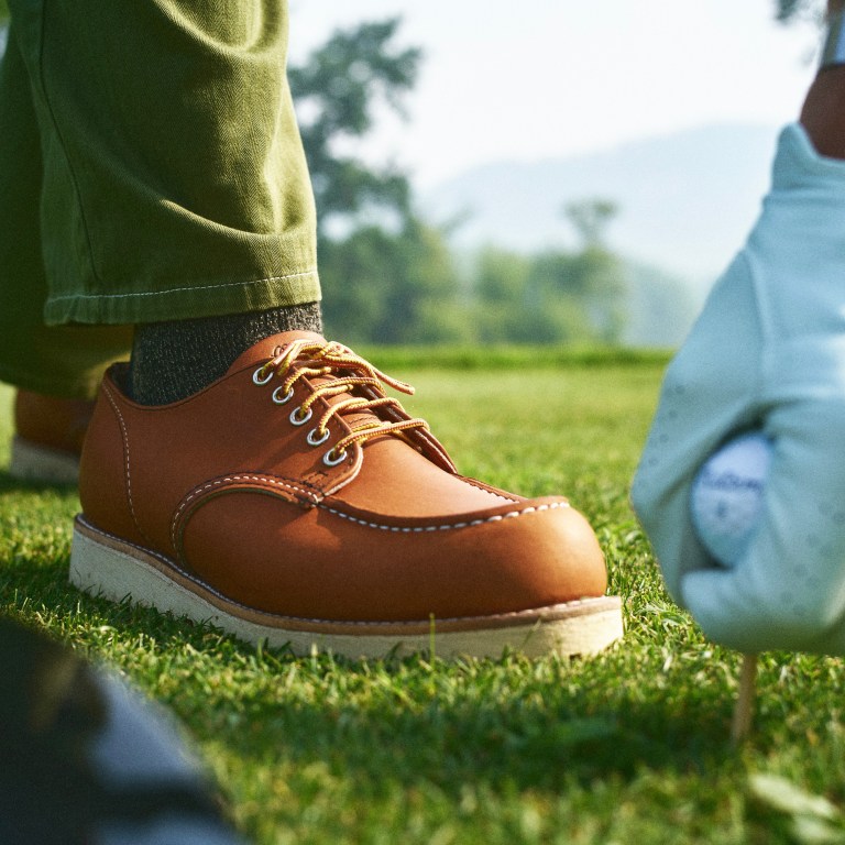 Malbon X Red Wing's new Moc Oxord, designed for golf, is shown on a man's feet alongside his hand placing a golf ball on a tee on a green