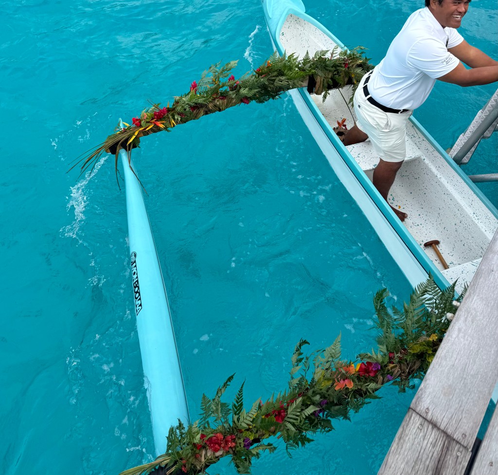 A tight crop of an overhead angled of a small Tahitian canoe with a man in the far right corner of the boat as it floats on crystal blue water.