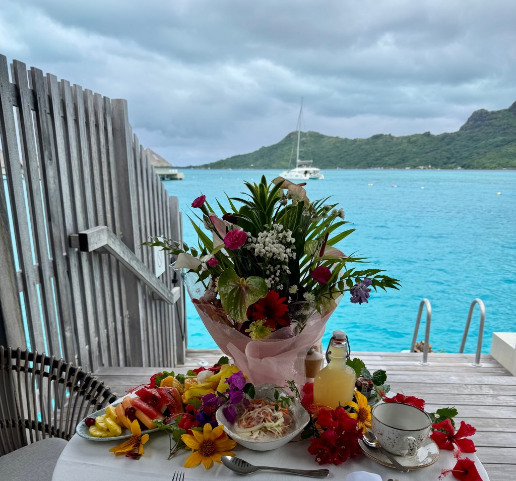 A table of colorful flowers and plated tropical dishes, looking out onto the lagoon beyond.