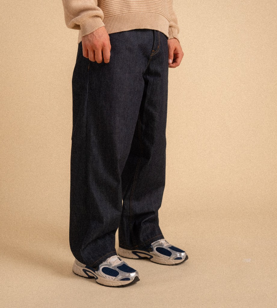 A model wears a pair of 1X dark denim jeans with a tan sweater, standing on a tan seamless background.