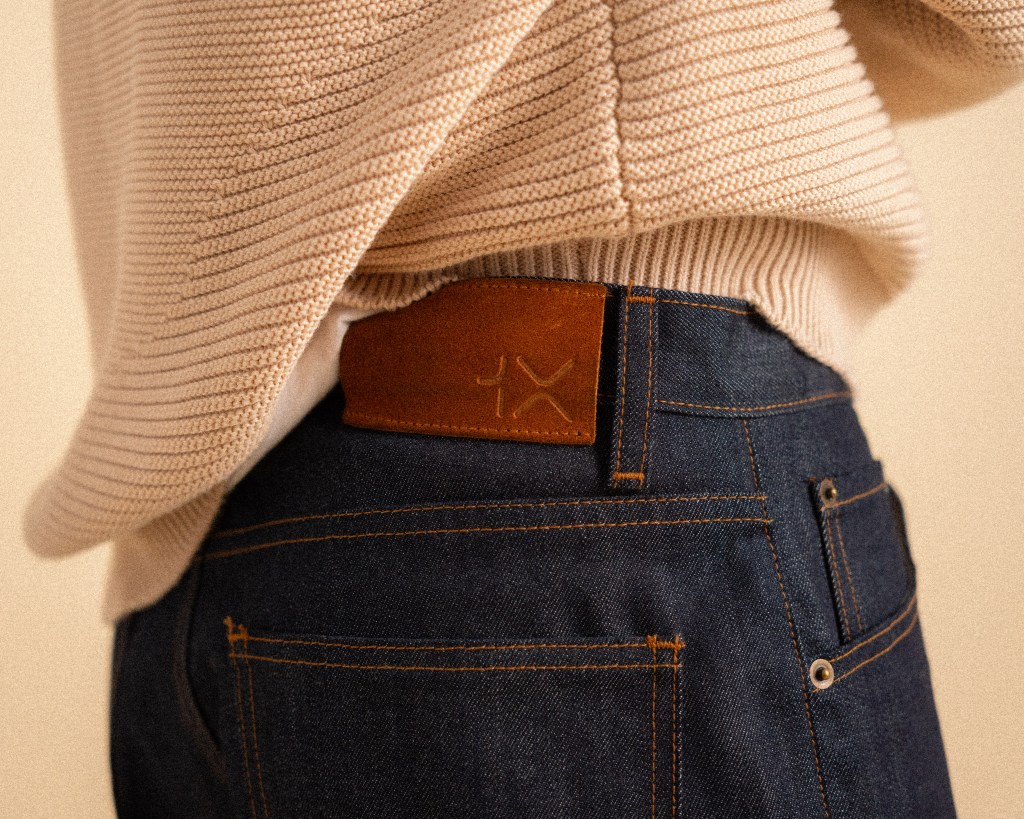 A detail of a model wearing a pair of 1X dark denim jeans and a light tan knit sweater.