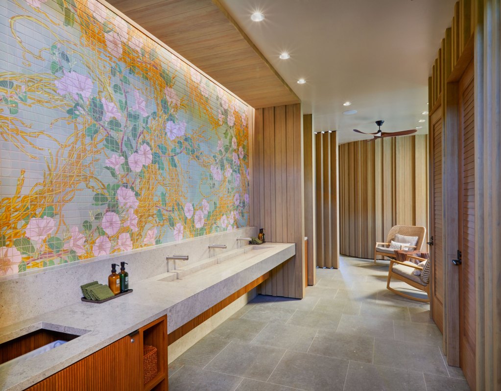 A wide painted mural above the long sinks in the women's spa washroom with LED lighting above it, with views of light wood walls and two rocking chairs in the background.