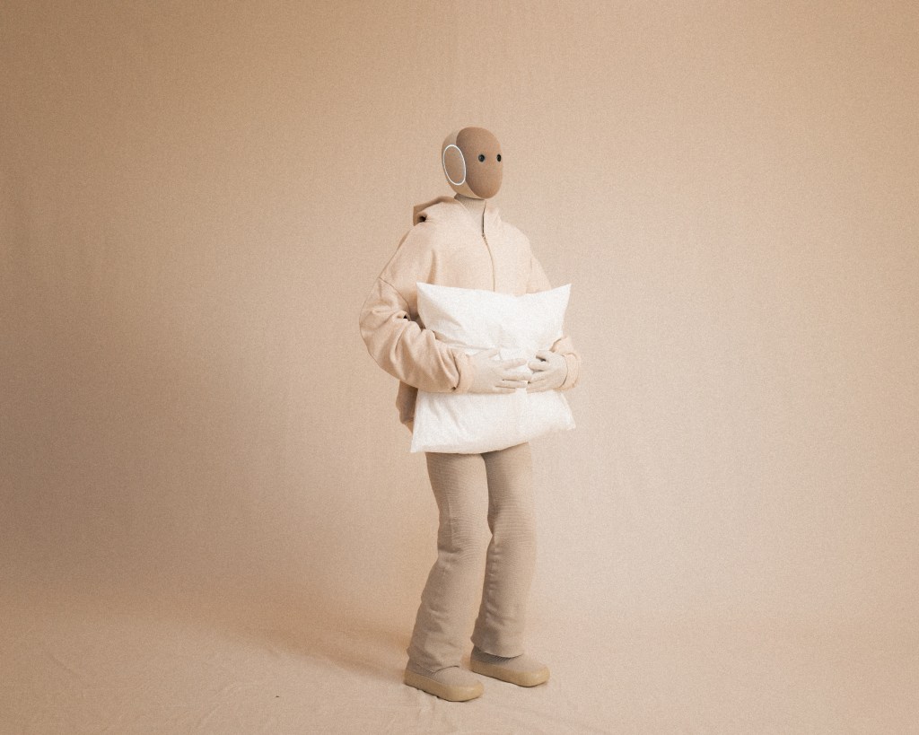 A beige humanoid robot in a soft knit outfit, holding a pillow, while standing on a light tan seamless background.