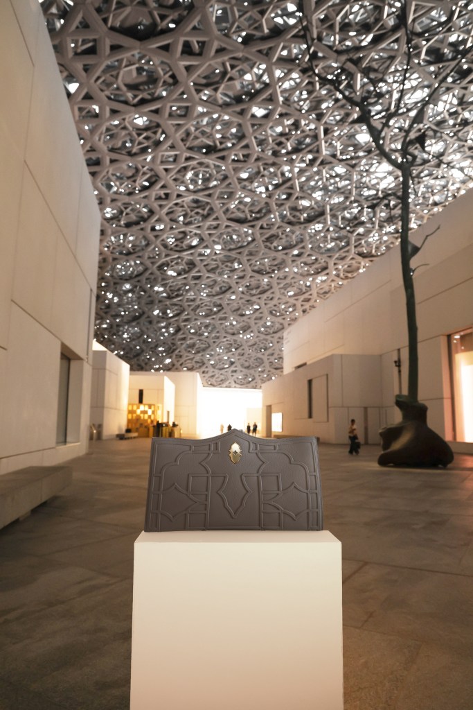 The gray Dellaluna Louvre Abu Dhabi hangbag sits on a pedestal in the museum with an ornate, lattice-like gray dome above it.