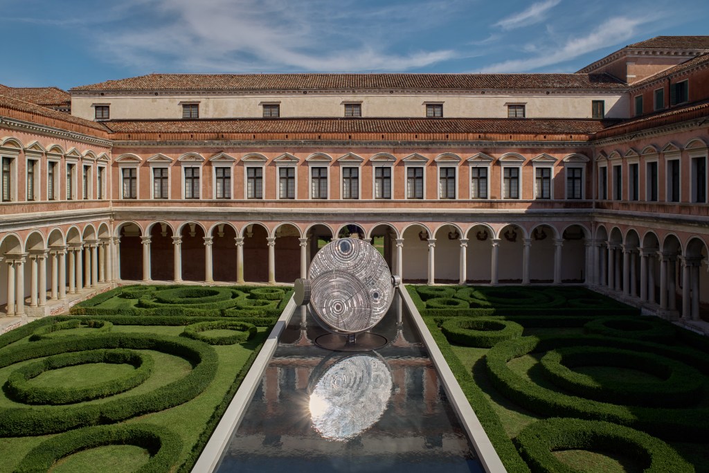 A large stone building with a lush, green yard and a large scale mirrored sculpture in front.