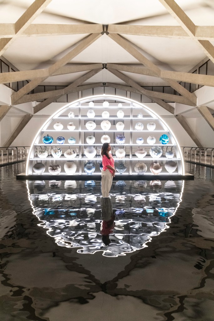 A woman wearing a pink top and cream pants stands in front of a large semi-circular installation while standing in shallow water while the artwork reflects to create a full circle.