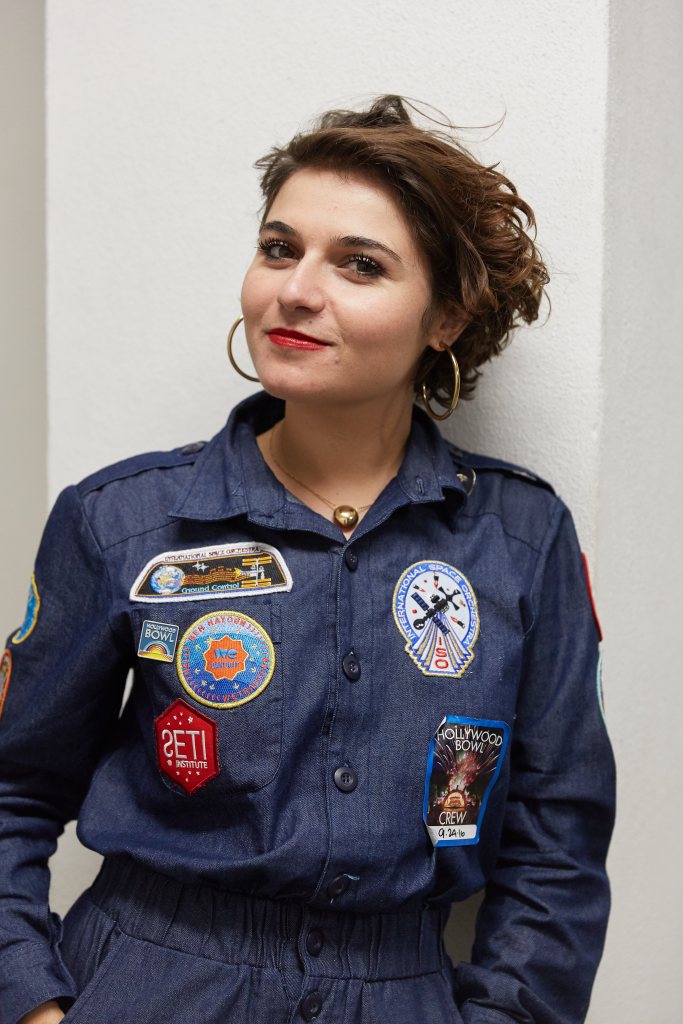 Nelly Ben Hayoun-Stéphanian standing in front of a while wall wearing a navy blue jumpsuit covered in astronaut-like patches.