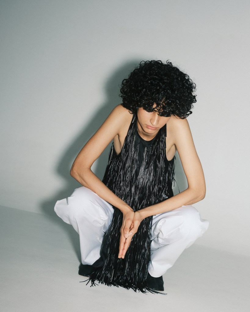 A model wearing a long black fringed top and white trousers squats and looks down at the white ground.