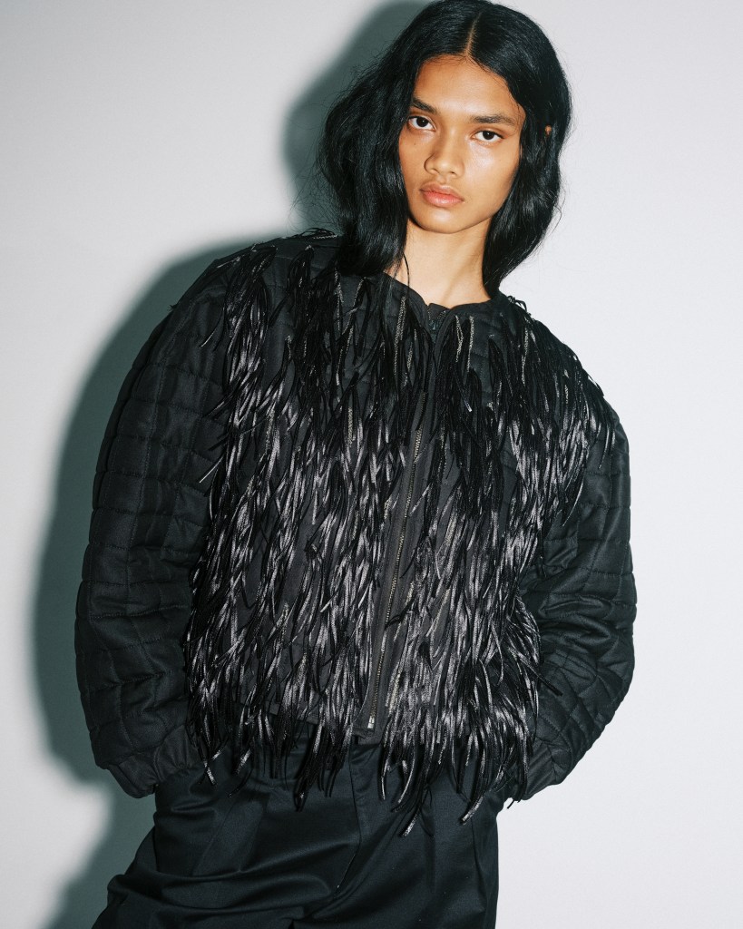 A model wearing a black silk and fringe bomber jacket, looking directly at the camera.  