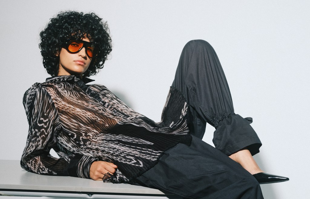A model wearing a sheer printed top and black trousers that tie at the ankle leans back on a white surface.