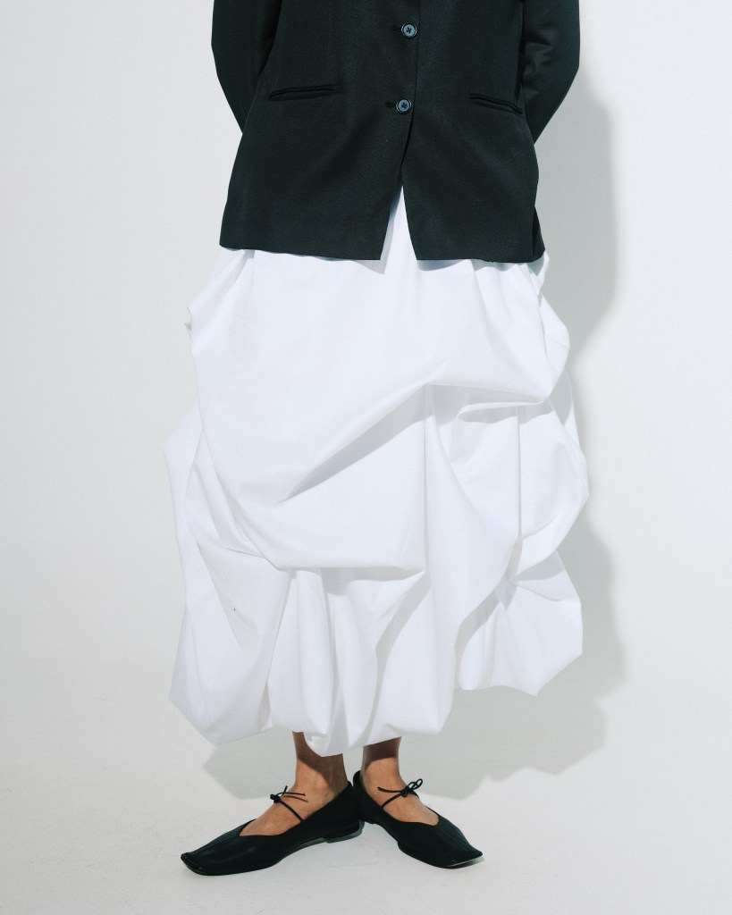 A shot of the bottom half of a model wearing a sculptural long white skirt, a black blazer and pointy black flats.