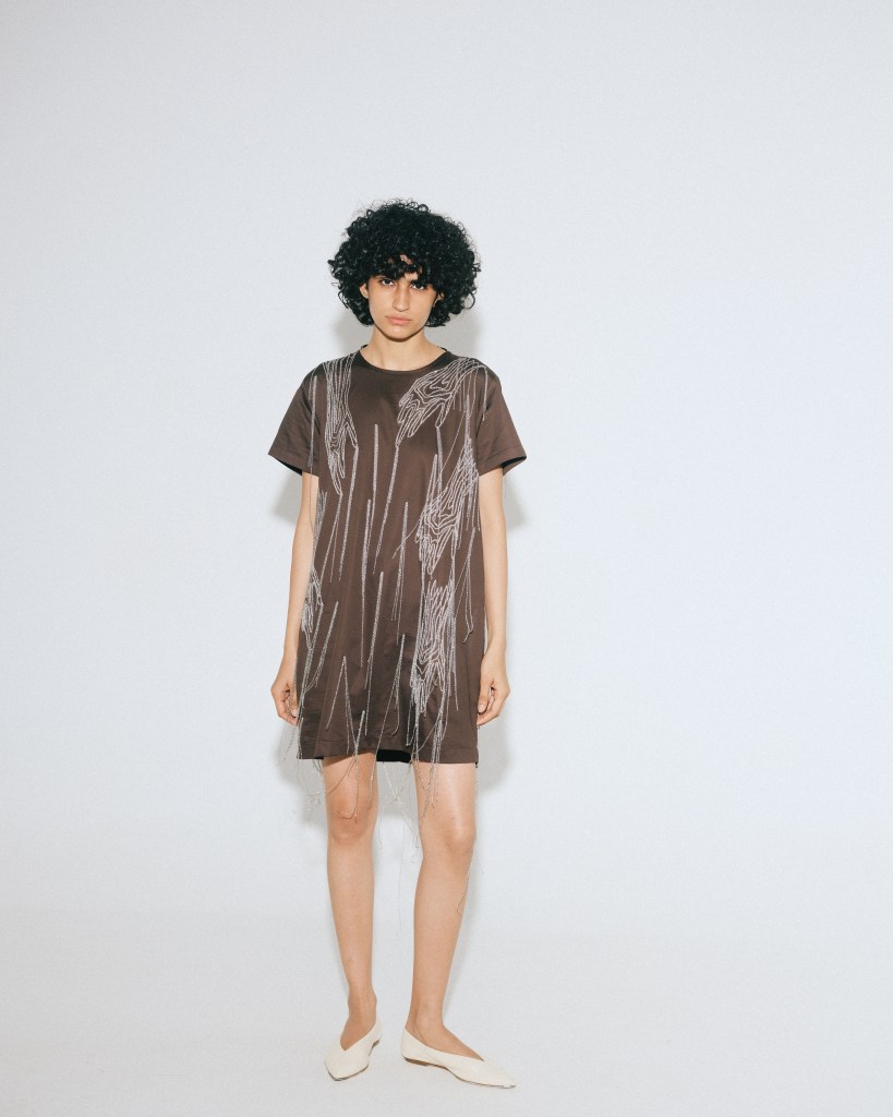 A model looks at the camera while wearing a brown t-shirt dress embroidered with a contrasting hand design.