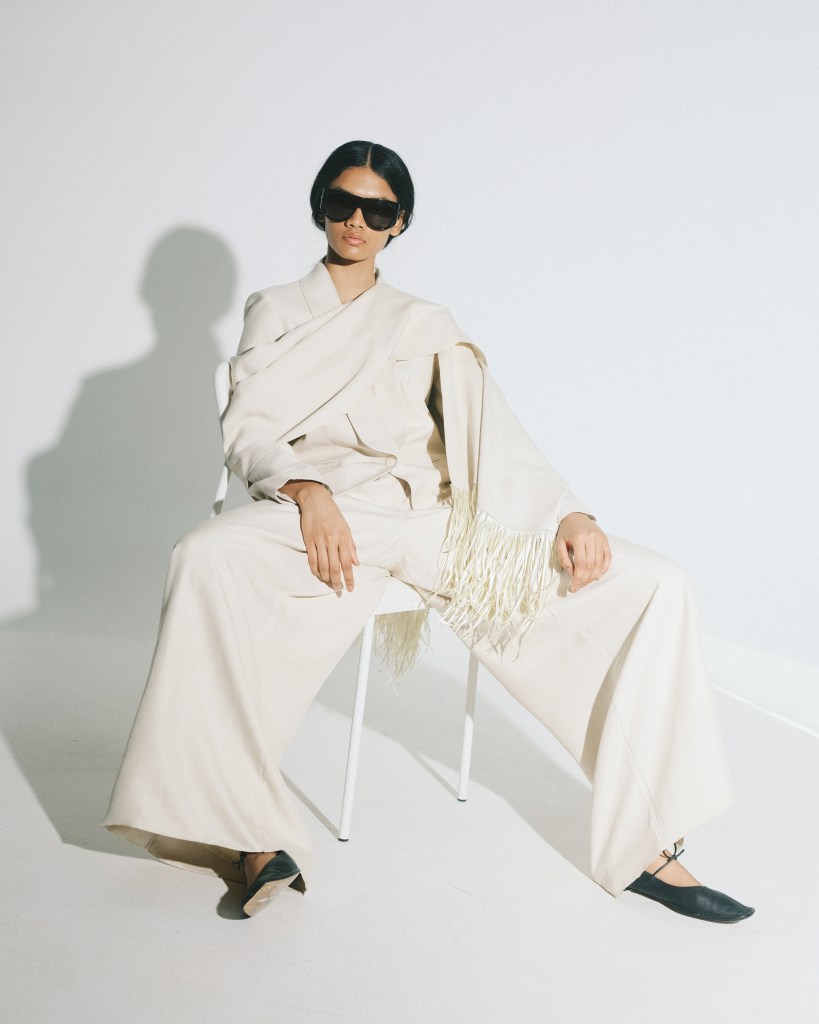 A model sits on a chair in a white room with a cream oversized suit and black sunglasses.