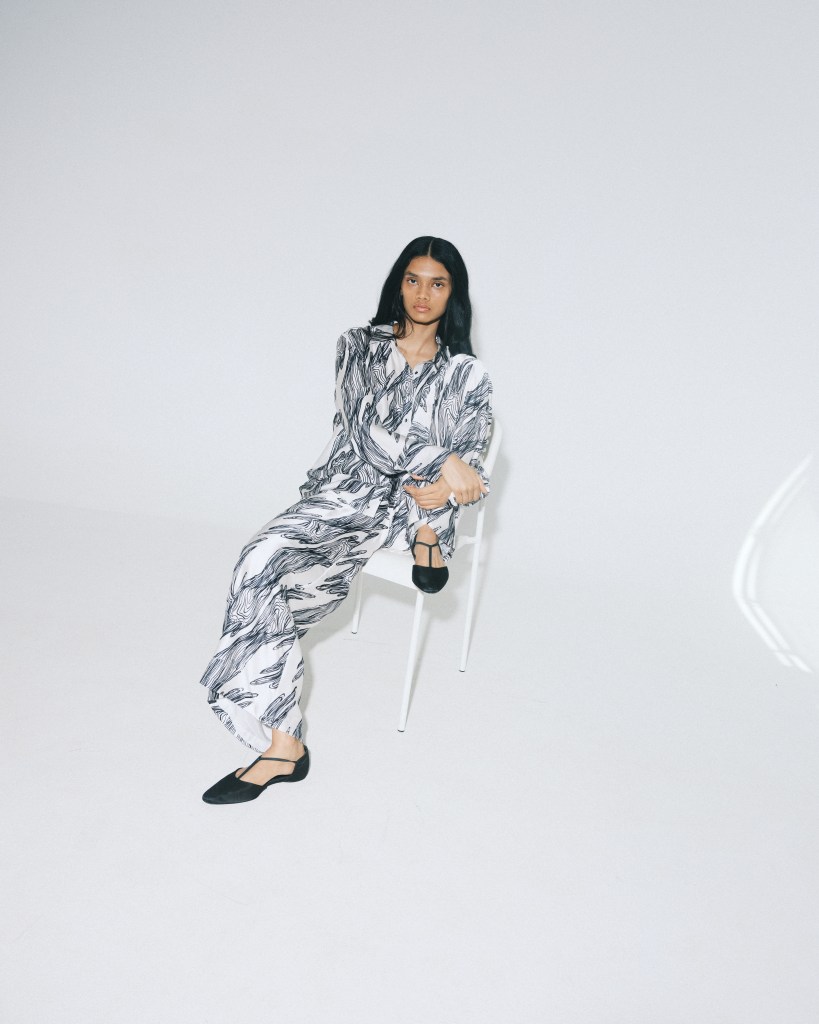 A model sits on a clear acrylic chair, wearing a  white top and wide leg pant covered in a black abstract pattern.
