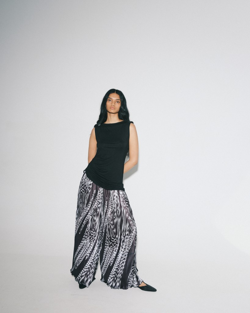 A model wearing a black sleeveless top and wide leg patterned pants stands with one leg turned out, looking at the camera.