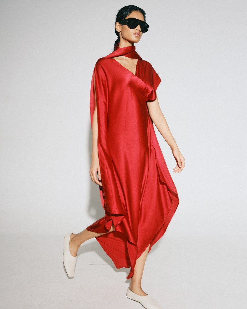 A model walks to the right of frame wearing sunglasses and a flowy red silk dress.