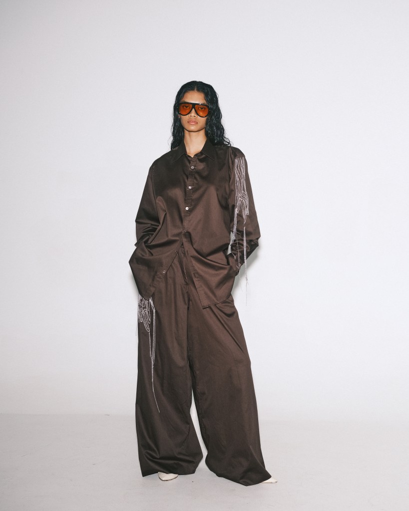 A model wearing sunglasses wear a button down brown top and wide leg brown trousers, with a hand embroidery detail on the top.