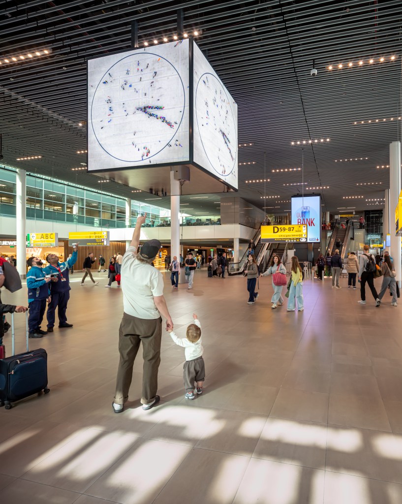 A father and baby, along with other passersby in the Amsterdam airport, point up at a large white cubic clock attached to the ceiling in the Amsterdam airport, with human figures creating the minute and hour hands. 