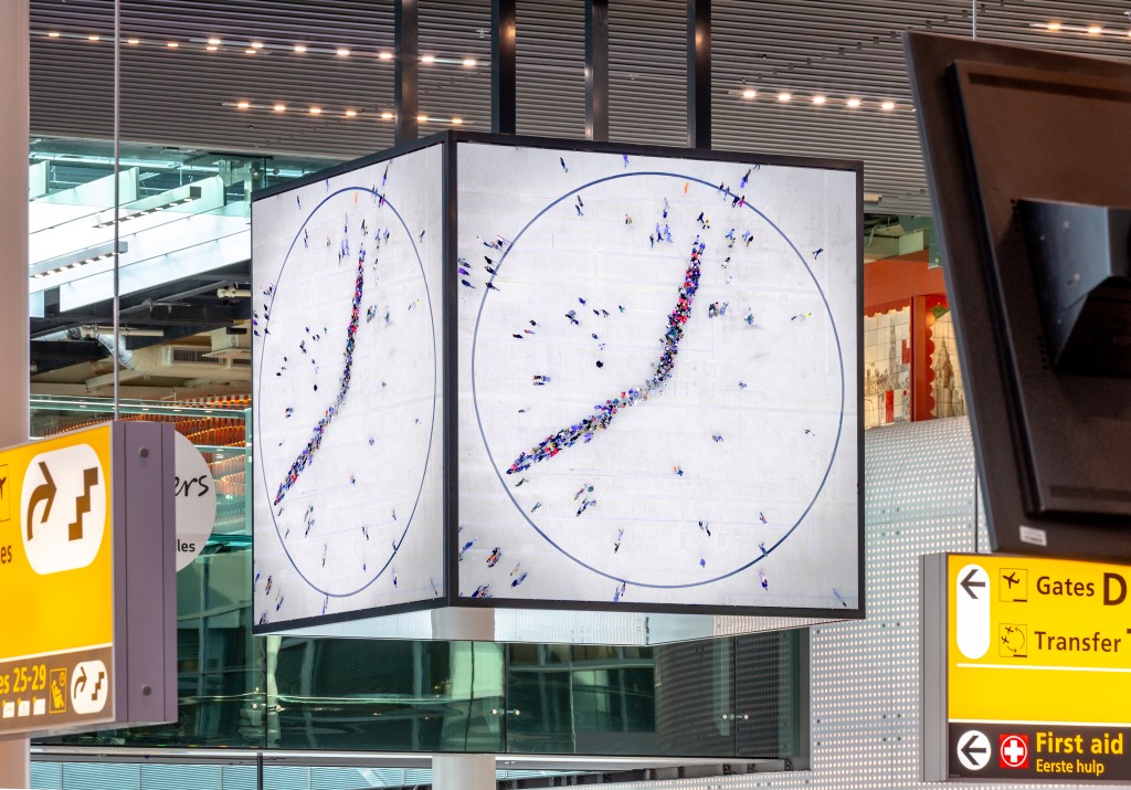 A large white cubic clock attached to the ceiling in the Amsterdam airport, with human figures creating the minute and hour hands.