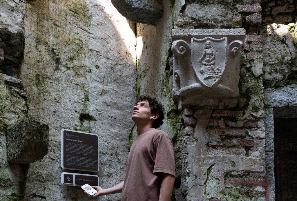 A man exploring the Castelli di Cannero with a phone in hand and headphones in his ears, as he looks up, standing in front of an ancient wall.