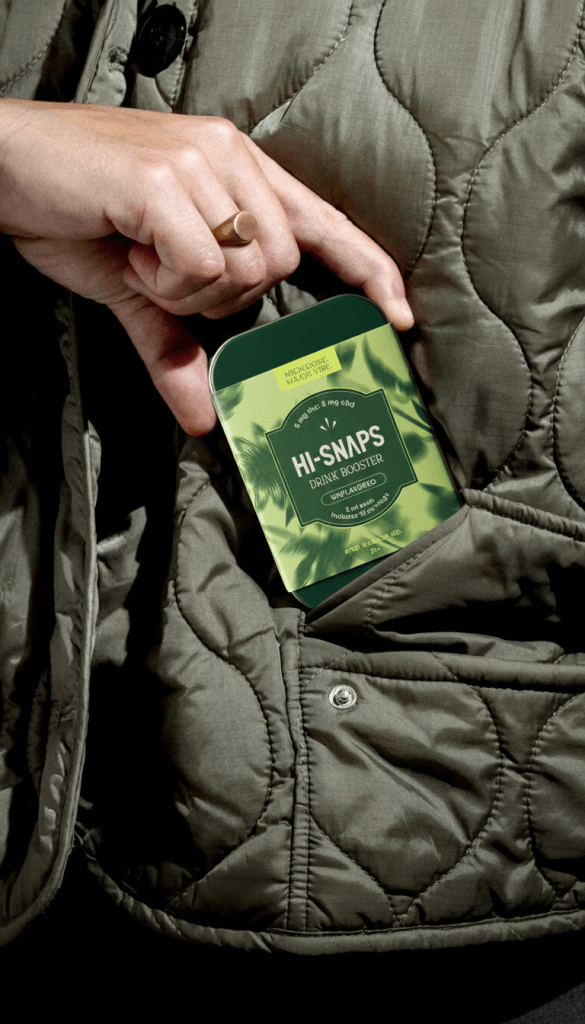 A hand holding a tin of  unflavored Hi-Snaps over the pocket of an army green quilted jacket.