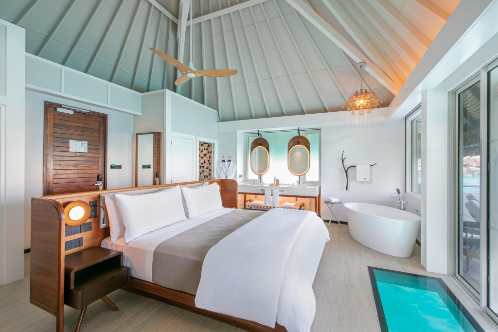 The interior view of a vaulted ceiling bungalow with a glass panel on the floor revealing crystal blue lagoon water below.
