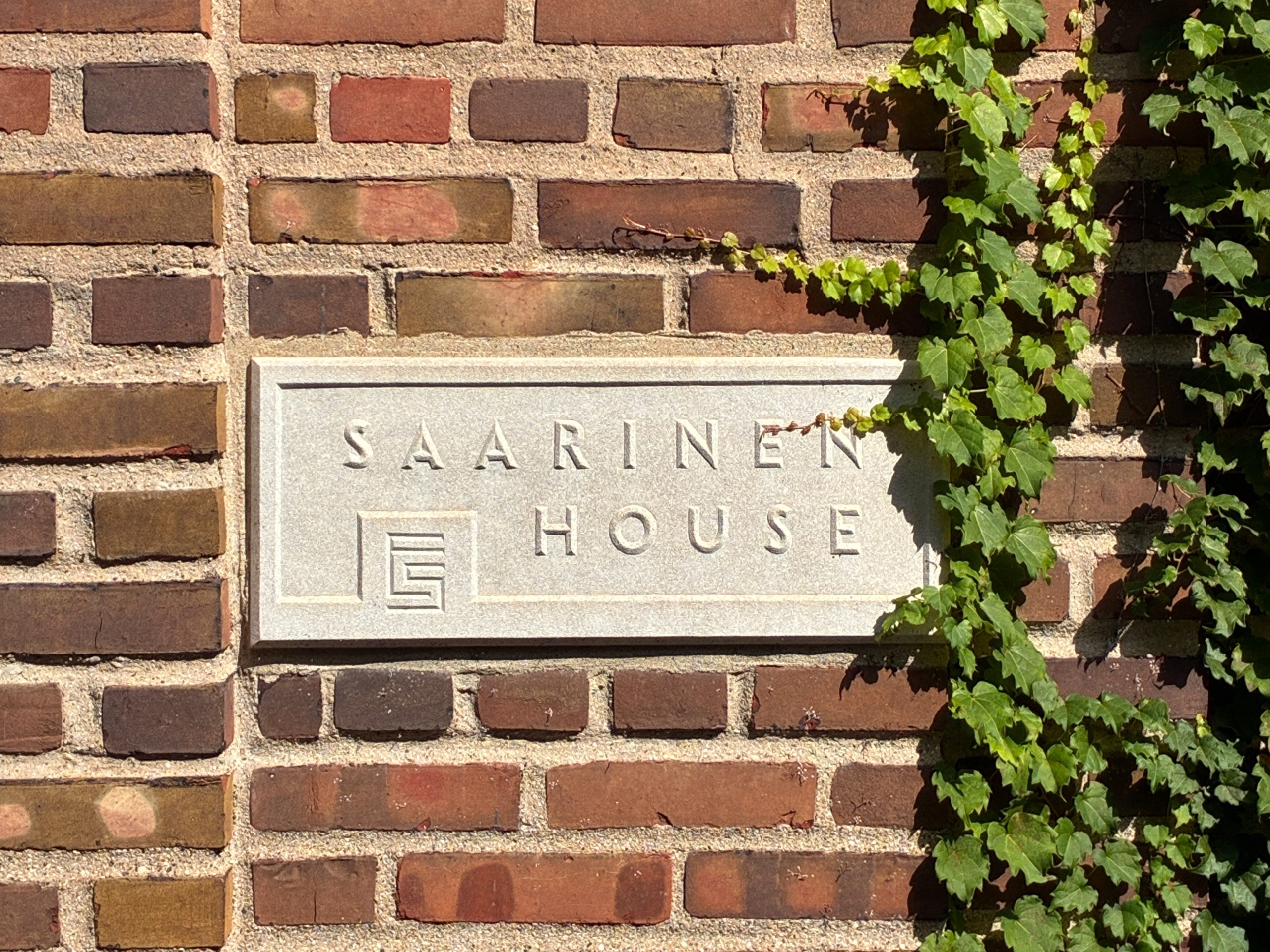 A sign that says "Saarinen House" on a brick wall with ivy creeping over it.