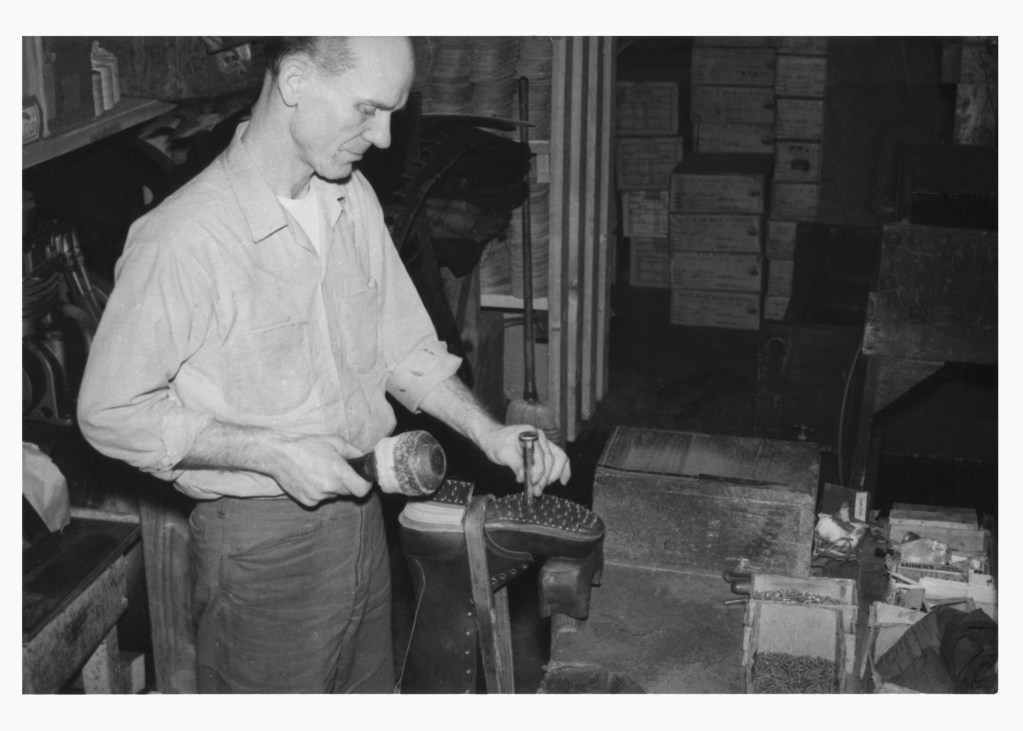 A black and white archival photograph of a shoemaker hammering the sole onto a pair of leather boots.