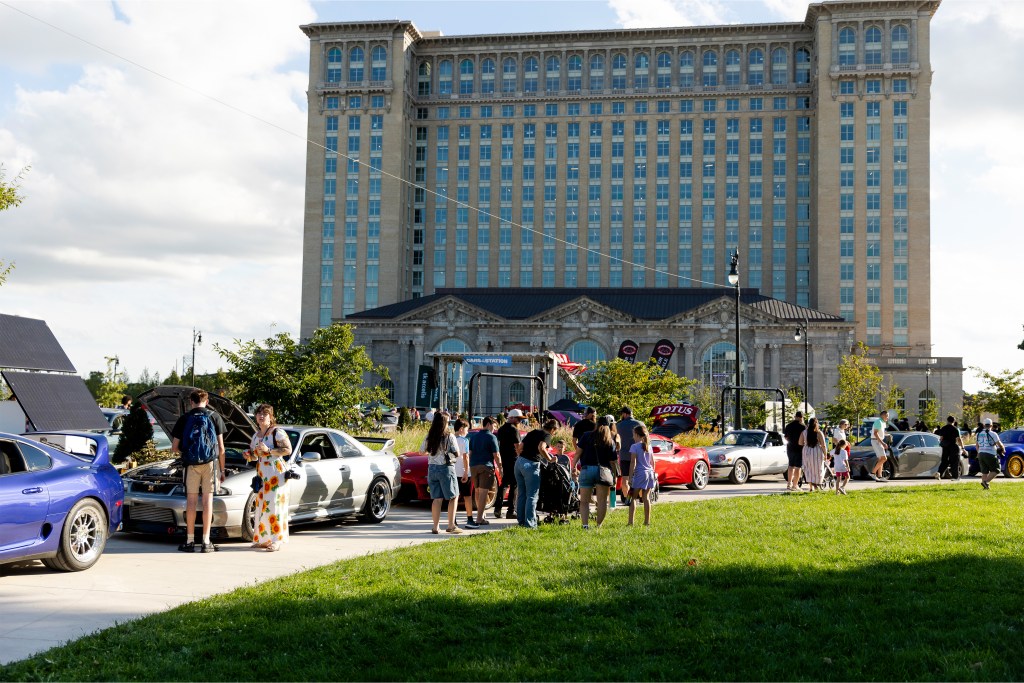 A group of people standing near a variety of colorful cars with Michigan Central in the background.