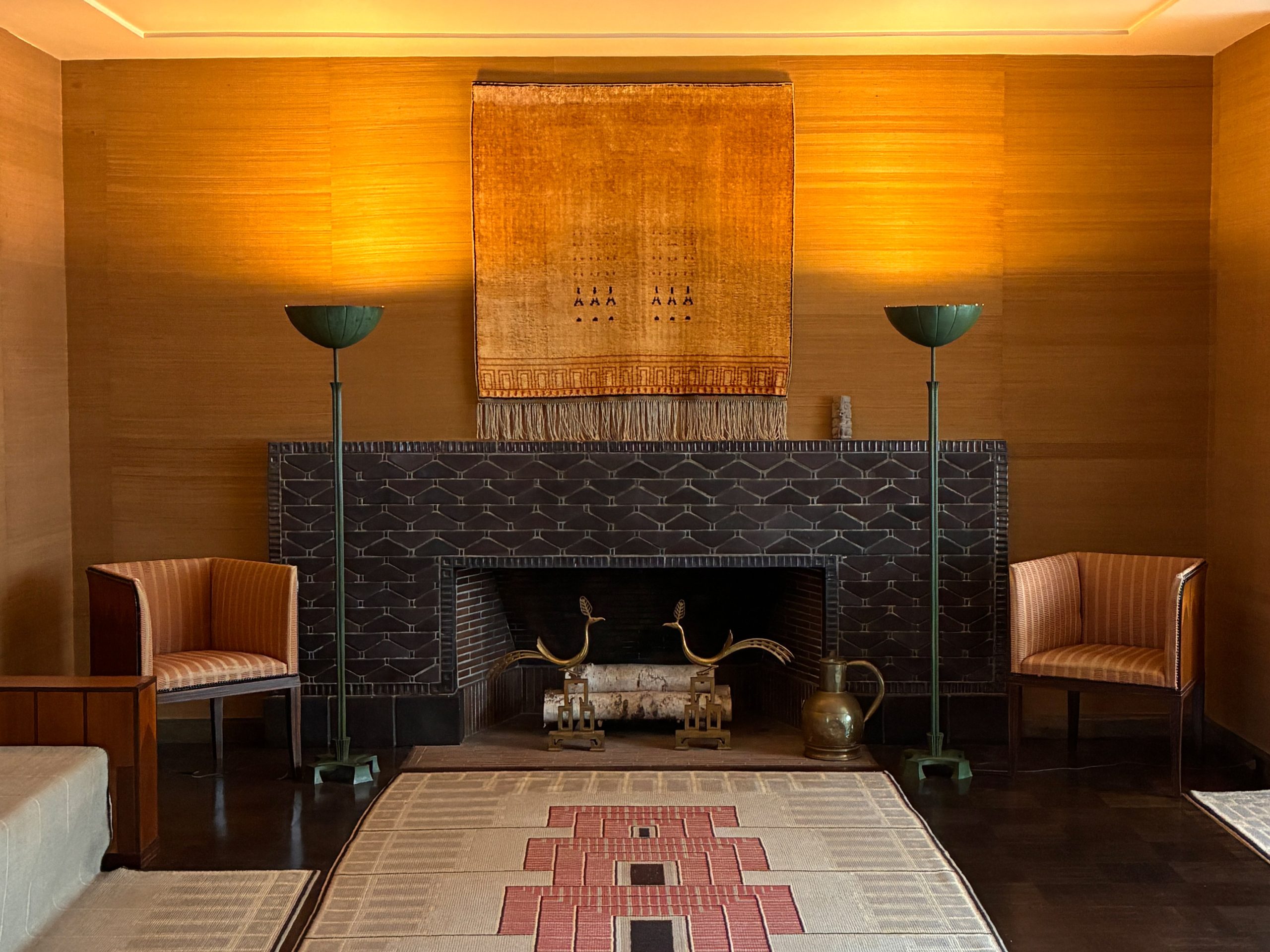 A living room view looking straight onto the fireplace and wood paneled wall with a cream and red red rug on the wood floor in front of it.