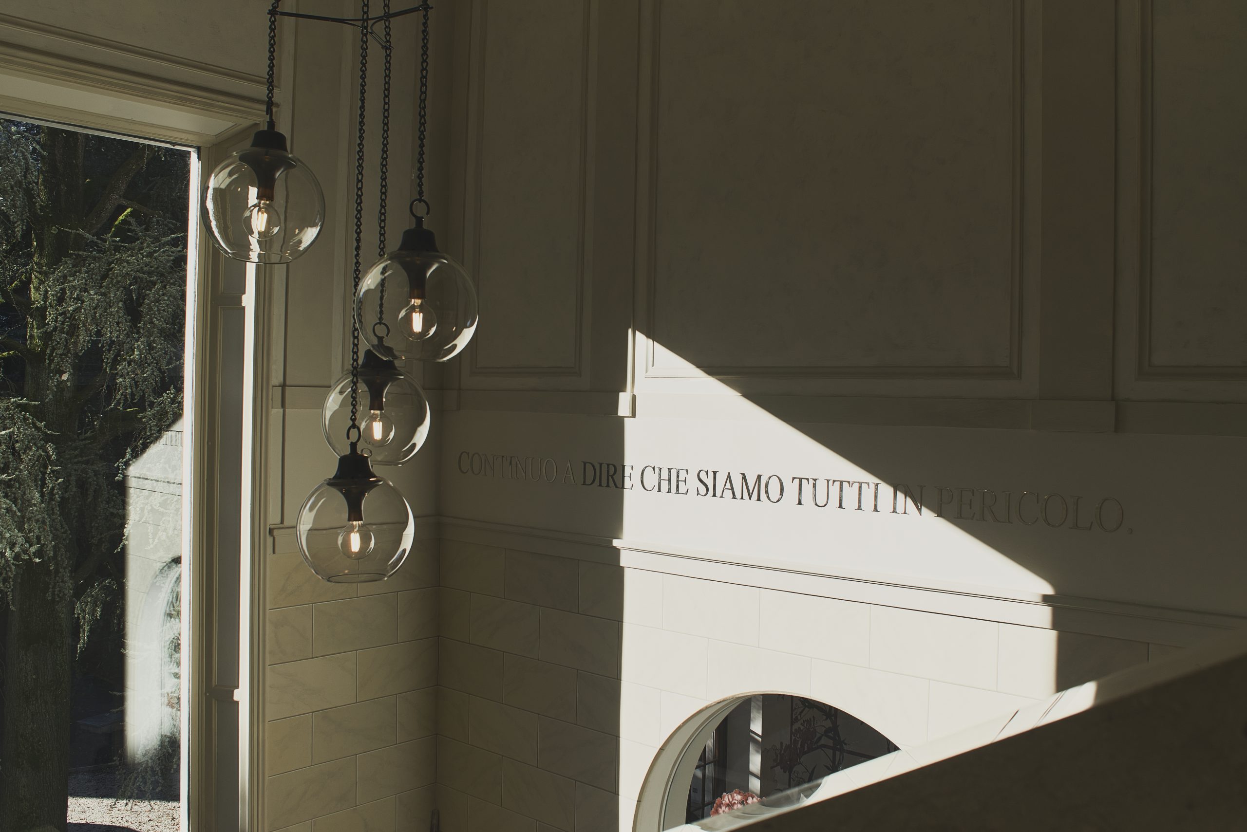 A marble stairwell with words inscribed on the wall and a large window illumating the wall and a hanging glass light fixture.