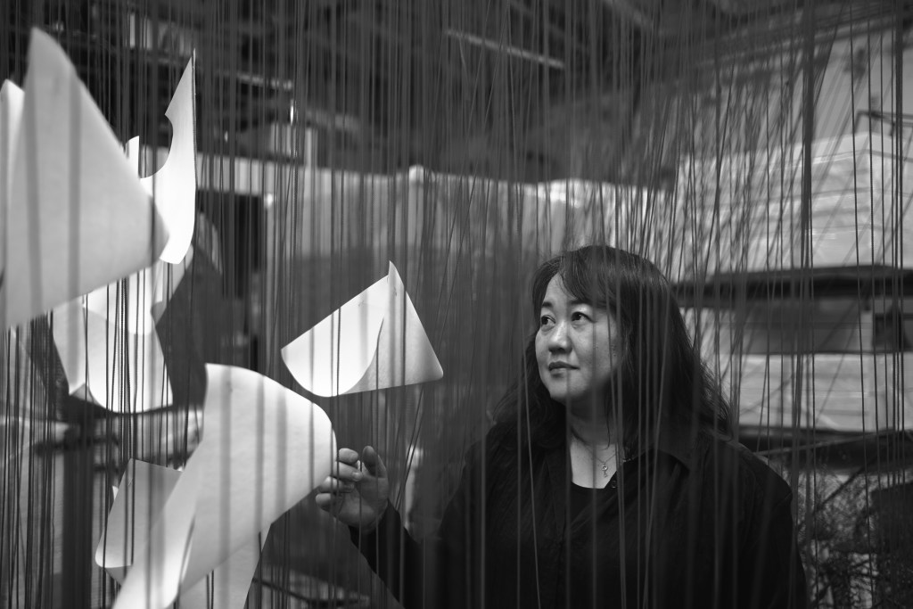 A black and white photo of Chiharu Shiota standing amongst her tactile, thread-based sculptural artworks.