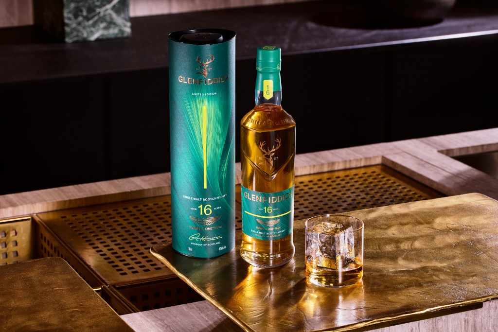 A bottle of Glenfiddich x Aston Martin Formula One collab, sitting on a wooden bar with a rocks glass in front of it.