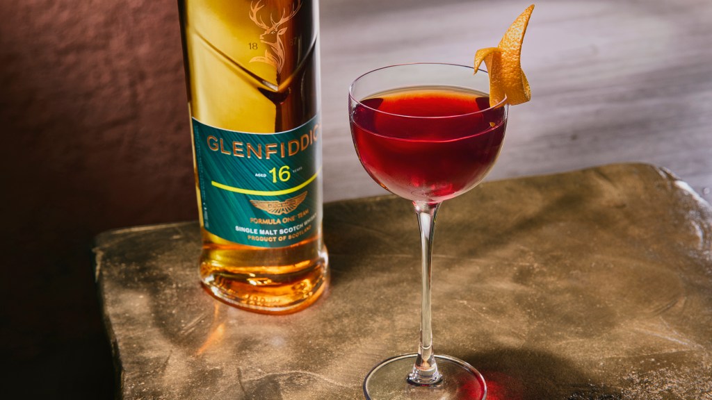 A detail of a cocktail with an orange slice sitting in front of a bottle of Glenfiddich x Aston Martin Formula One Team scotch.