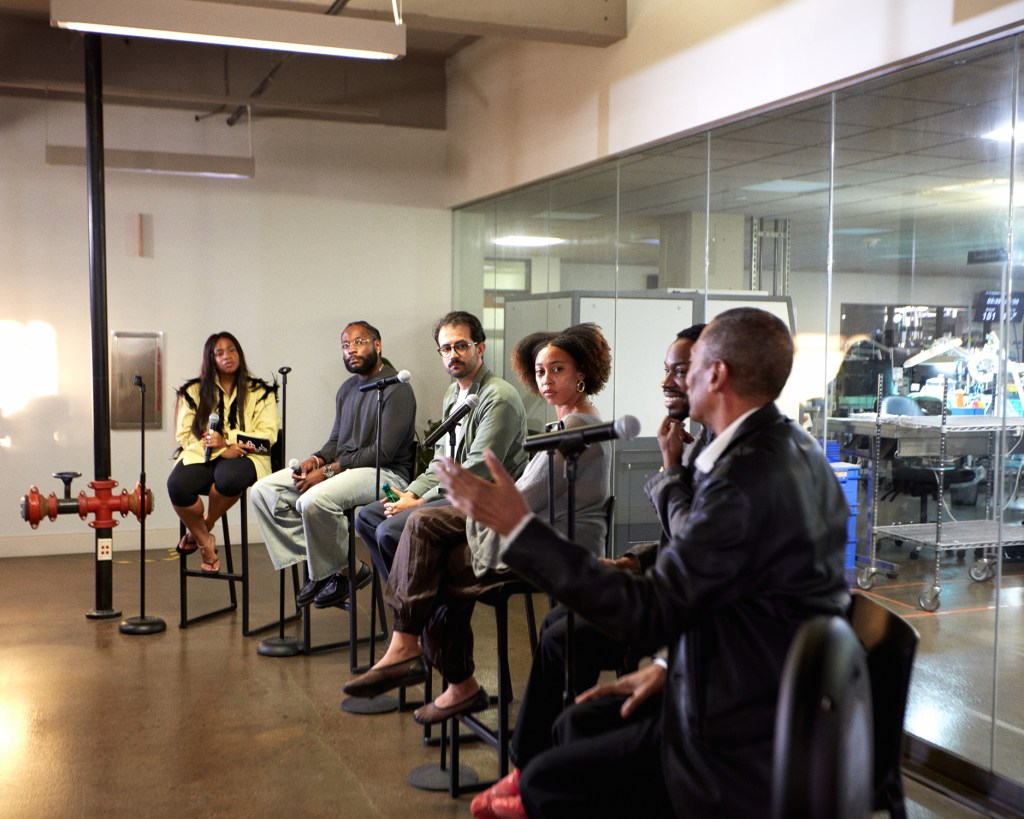 A group of people sit in a row on stools in front of a glass-walled room.