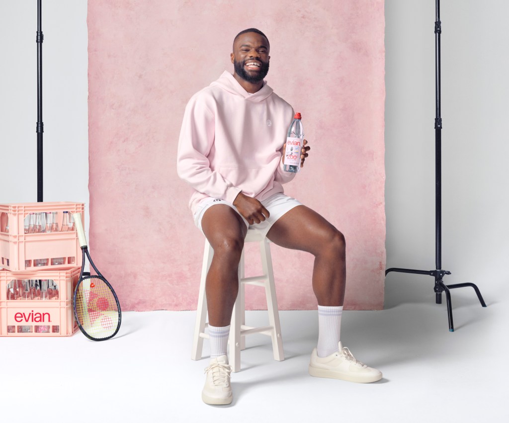 Frances Tiafoe sits on a white stool wearing a light pink sweatshirt and white shorts in front of a light pink seamless background in a photo studio, holding an evian bottle.