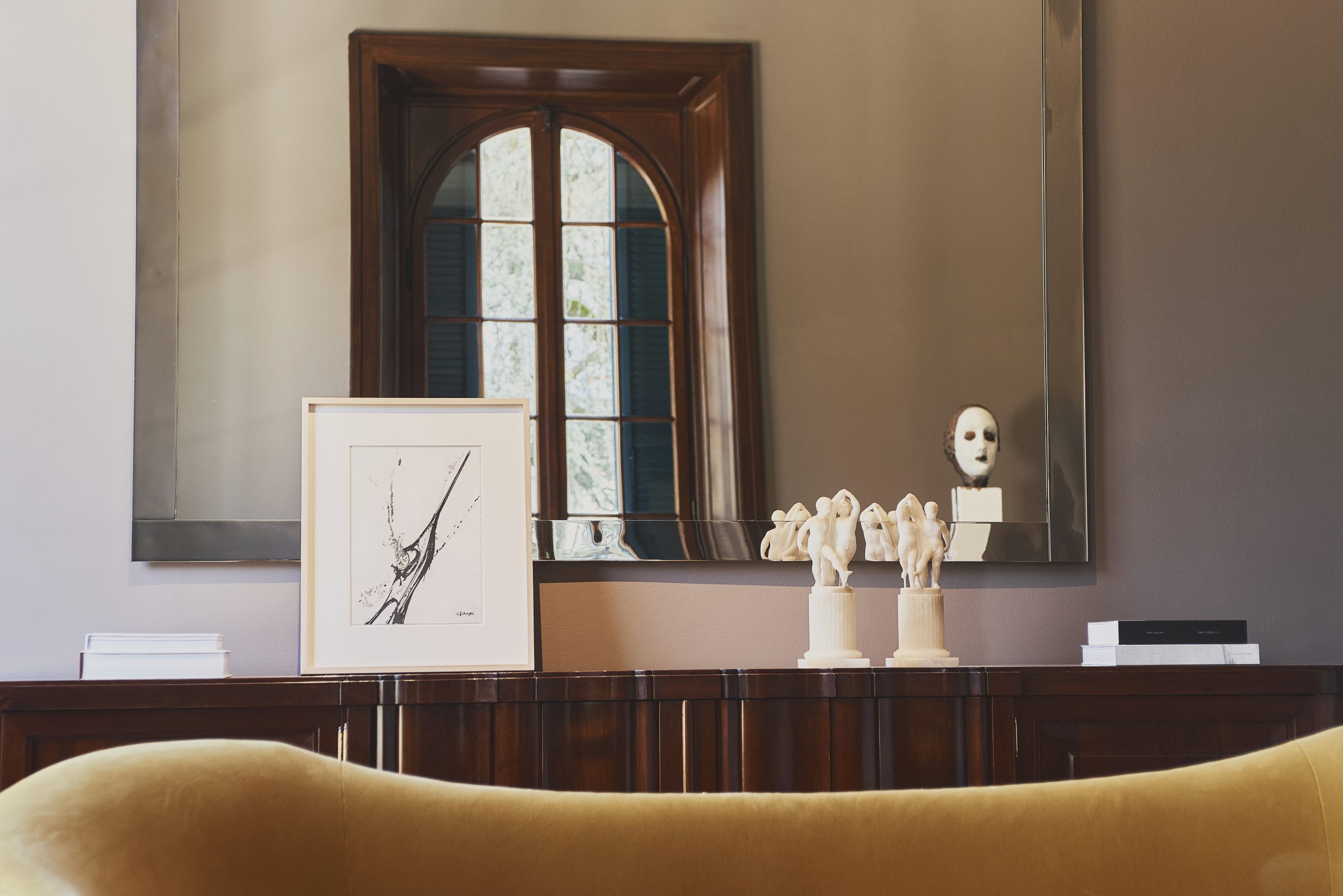 A wide wooden table with artworks displayed on it and a tall arched window behind it.