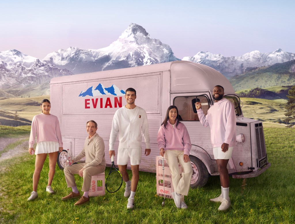 A group of athletes and artists including Frances Tiafoe, Carlos Alcaraz and Jeff Koons standing in front of a light pink evian bus with an image of the French Alps behind them.