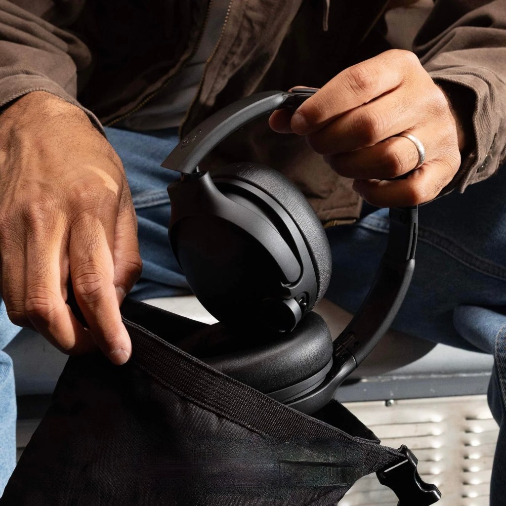 A man holds a folded pair of Skull Candy's Crusher 1080 headphones while seated