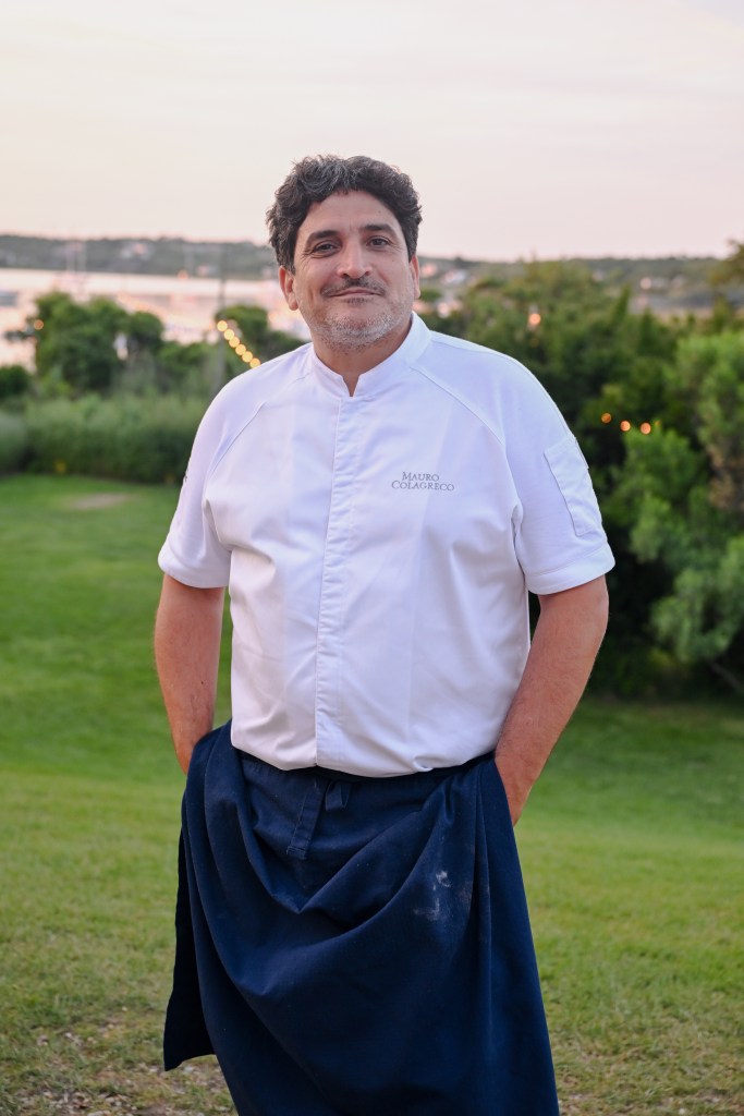 Mauro Colagreco stands with a chef shirt an navy apron, hands in his pockets, with a smile on his face and an early sunset behind him.