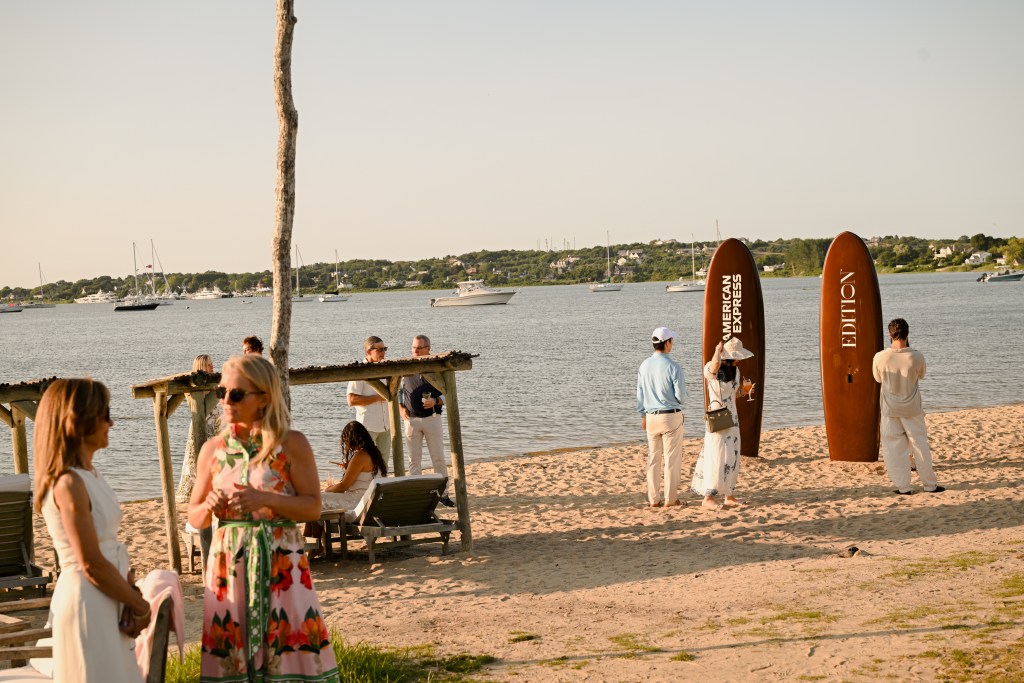 A beach with calm water and people milling around, and two brown surfboards with white signage sticking up vertically. 