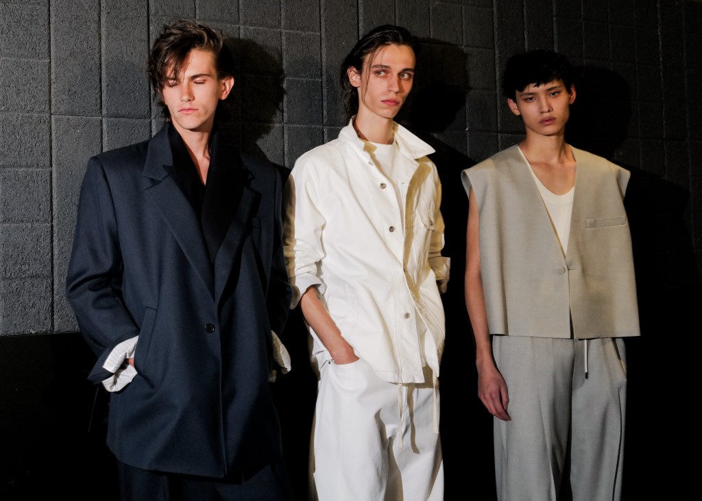 A backstage scene of three models wearing neutral-colored suits, standing against a dark wall.
