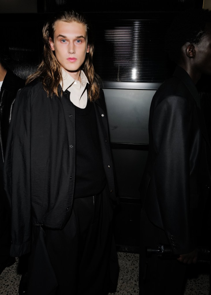A male model with long blonde hair and wearing a white collared shirt under a black sweater and jacket gazes into the camera.
